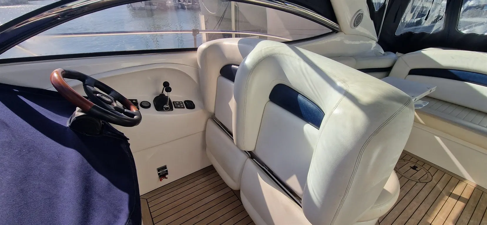 Spacious boat interior featuring a steering wheel, control panel, and leather seats with wooden flooring, beside the water.